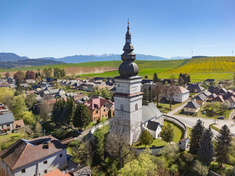 Aerial view of the historic St. Matthew's Church tower and traditional houses surrounded by green fields and distant mountains under a clear blue sky in Partizanska Lupca, Zilina Region, Slovakia.