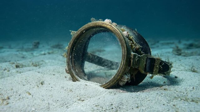 Barnacle-covered diving mask on sandy seabed, static shot, underwater, for marine exploration.