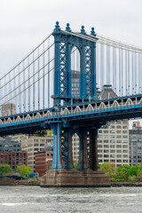Naklejka premium Blue steel tower of Manhattan Bridge over East River. Architectural detail of Manhattan Bridge suspension tower with brick buildings and river water in New York.