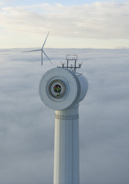 Netherlands - 26 November 2025: Aerial view of workers inside a wind turbine nacelle rising above a thick layer of clouds under a soft sky.