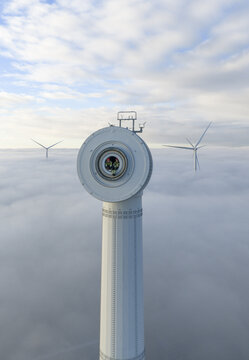 Netherlands - 26 November 2025: Aerial view of a wind turbine nacelle with workers inside rising above a thick layer of fog under a blue sky.