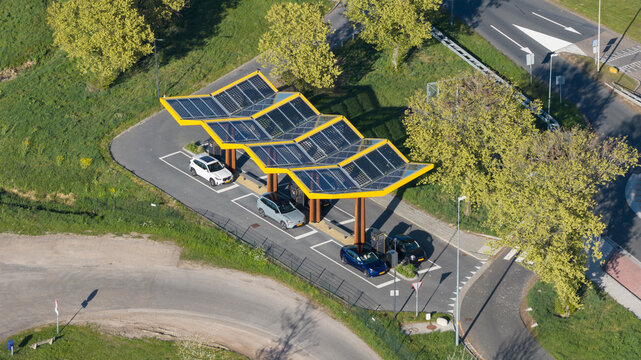 Bant, Netherlands - 23 April 2026: Aerial view of Fastned Charging Station with its yellow solar-paneled roof, electric vehicles parked, and surrounding green trees under bright daylight.