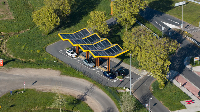 Bant, Netherlands - 23 April 2026: Aerial view of the Fastned Charging Station featuring its iconic yellow solar panel roof with electric cars parked in charging bays surrounded by lush green trees.