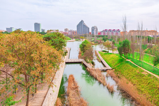 Cabecera Park in Valencia with tranquil artificial lake surrounded by lush green slopes and modern apartment buildings. Wooden walkways bridge calm water, offering peaceful urban escape for strollers