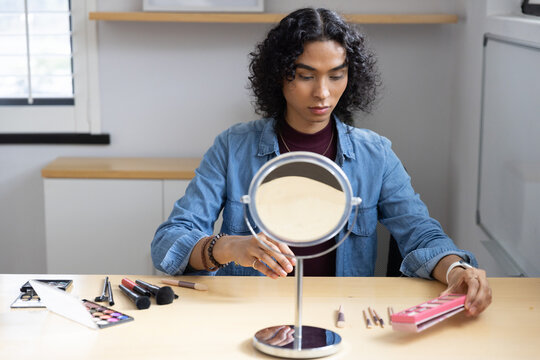 Nonbinary person in denim shirt and maroon top arranging makeup at home with mirror, brush, palette