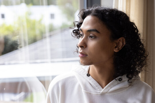 Non-binary person gazing toward right through large window at home, wearing white hoodie
