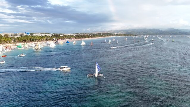 Malay, Philippines - 02 May 2026: Aerial view of Boracay Island with numerous sailboats on the turquoise water along the white sand beach under a cloudy sky with a faint rainbow.