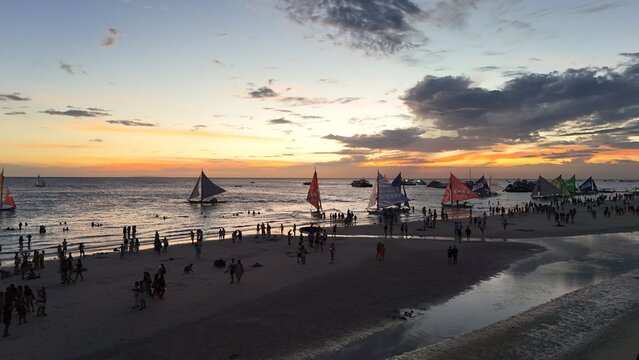 Malay, Philippines - 02 May 2026: Aerial view of Boracay Island beach at sunset with numerous traditional paraw sailboats and people gathered on the white sand under a dramatic orange sky.