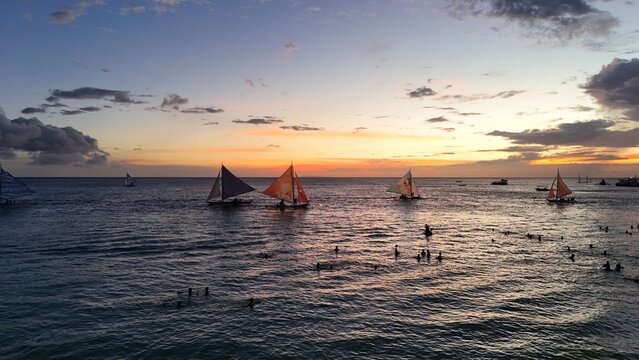 Malay, Philippines - 02 May 2026: Aerial view of Boracay Island with traditional paraw sailboats sailing on the calm sea during a vibrant sunset, with people swimming in the water.