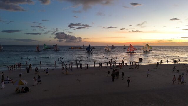 Malay, Philippines - 02 May 2026: Aerial view of Boracay Island featuring crowds of people on the white sand beach and traditional sailboats on the calm sea during a vibrant sunset.