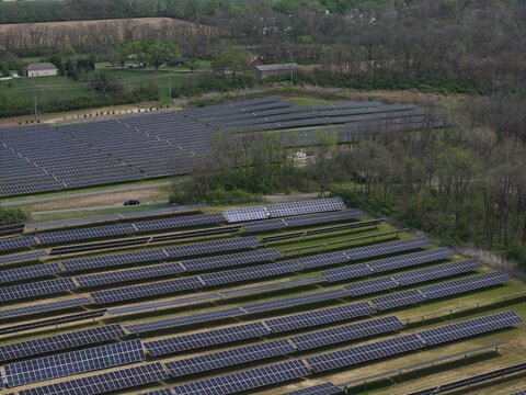Lebanon, United States - 12 April 2026: Aerial view of a large solar farm with rows of photovoltaic panels surrounded by lush green trees and rural fields under soft daylight.