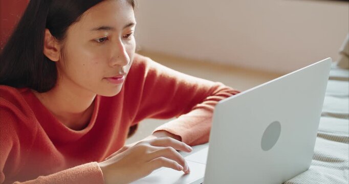 Asian teenager girl typing working with laptop on her bed, woman learning online pass internet at home, in Cozy Bedroom with Warm Morning Sunlight, Side View.