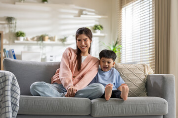 Obraz premium Happy asian mother and young child smile together on grey sofa in bright living room showing joyful family bonding and deep affection while enjoying peaceful morning sunlight at home relaxing