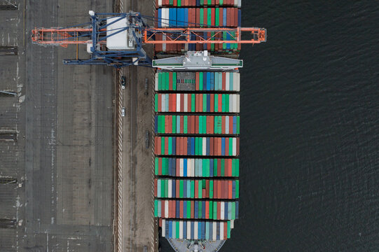 05 September 2019: Aerial view of a large container ship loaded with colorful cargo docked at a port terminal with an industrial gantry crane.