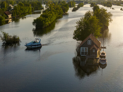 Breukelen, Netherlands - 25 April 2026: Aerial view of Scheendijk recreational area with a motorboat cruising past a wooden house on a small island surrounded by lush greenery and calm waters.