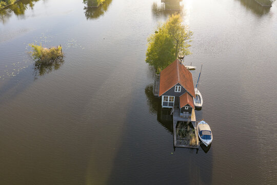 Breukelen, Netherlands - 25 April 2026: Aerial view of Scheendijk, featuring a traditional wooden house on a small island surrounded by calm water and moored boats during golden hour.