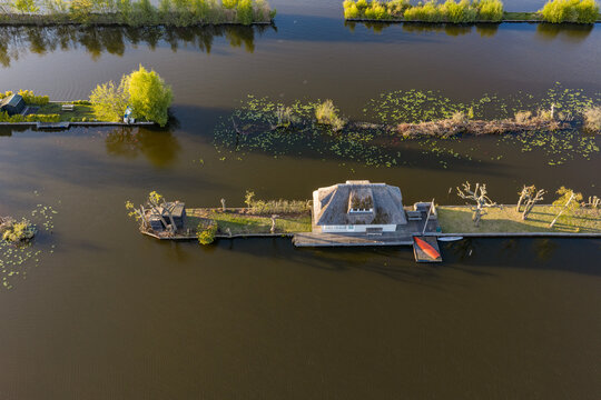 Breukelen, Netherlands - 25 April 2026: Aerial view of Scheendijk featuring a thatched-roof house on a narrow island surrounded by calm water and lily pads.