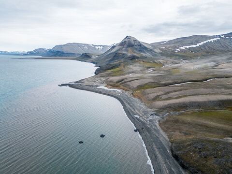 Svalbard and Jan Mayen - 17 July 2025: Aerial view of the rugged coastline with dark sand beaches, small expedition boats in the water, and snow-capped mountains under an overcast sky.