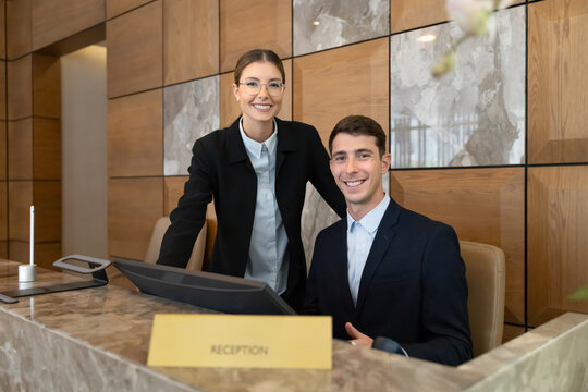 Portrait of professional hotel receptionists, colleagues posing behind front desk in modern hotel lobby, smiling, looking at camera, welcoming arriving guests. Hospitality services, tourism industry