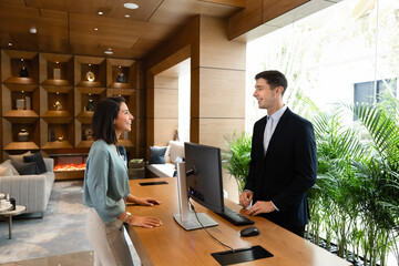 Fototapeta na wymiar Female guest standing at hotel reception desk speaking with male receptionist during check-in. Travel arrival process, hospitality service, customer interaction in modern hotel lobby, tourism concept
