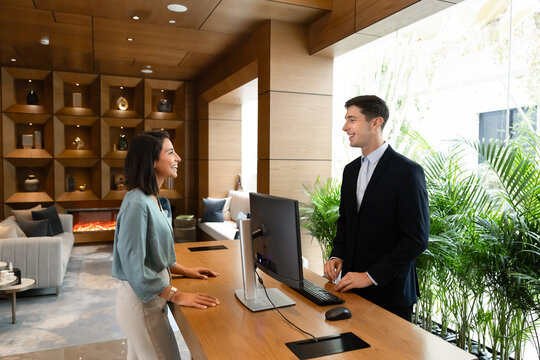 Female guest standing at hotel reception desk speaking with male receptionist during check-in. Travel arrival process, hospitality service, customer interaction in modern hotel lobby, tourism concept