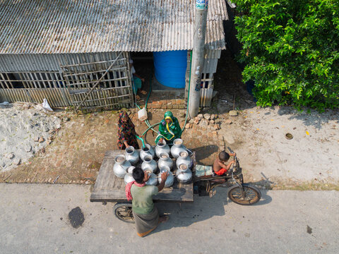 Satkhira, Bangladesh - 20 April 2026: Aerial view of people filling metal water jugs on a flatbed tricycle cart next to a corrugated metal building.