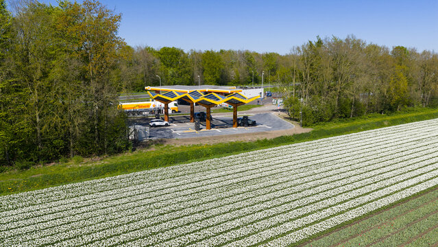 Bant, Netherlands - 21 April 2026: Aerial view of Fastned charging station with yellow solar roofs next to a blooming white tulip field and green trees.