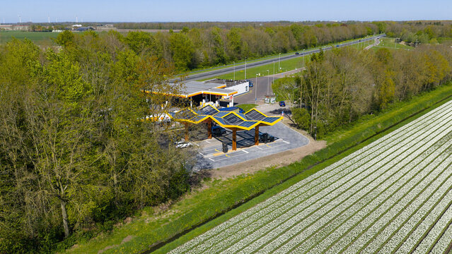 Bant, Netherlands - 21 April 2026: Aerial view of the Fastned Charging Station with its yellow solar roofs next to a highway and a blooming white tulip field.