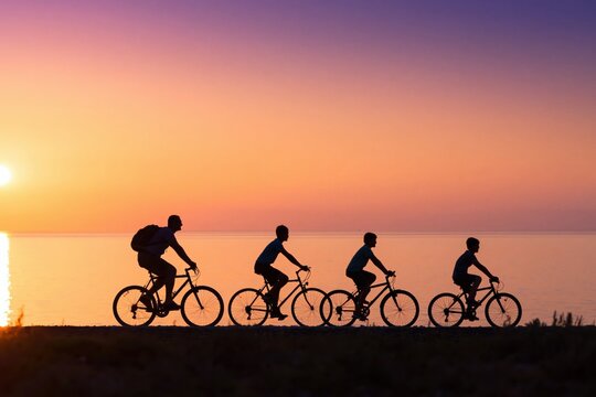 Family of four cycling along a coastal path at vibrant sunset, silhouettes against orange sky enjoying active, joyful seaside togetherness