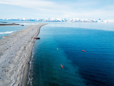 Svalbard and Jan Mayen - 29 June 2025: Aerial view of kayakers paddling in turquoise waters along a narrow pebble spit with snow-capped mountains in the background.