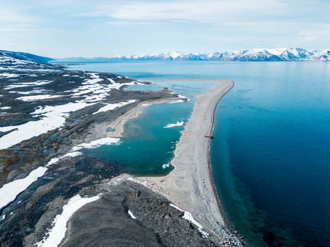 Svalbard and Jan Mayen - 29 June 2025: Aerial view of a long coastal sandbar separating turquoise lagoons from the deep blue sea with snow-capped mountains in the distance.