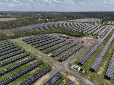 Lebanon, United States - 12 April 2026: Aerial view of a large solar farm with rows of photovoltaic panels installed on green fields surrounded by dense trees under a cloudy sky.