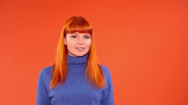 Person with orange hair talks and expresses different emotions in front of a bright orange background while wearing a blue sweater