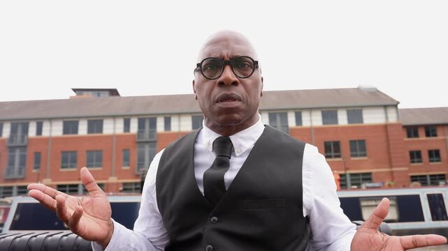 Man shares thoughts while seated near buildings and water, expressing feelings with hand gestures during daytime in a city
