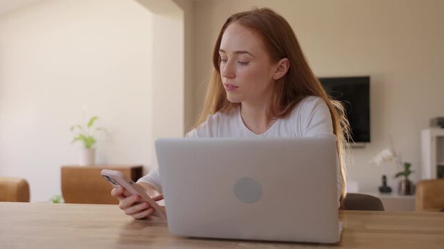 Woman working at home using laptop and phone in modern living room during afternoon