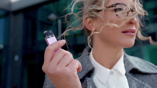 Woman holds vape outdoors while wind blows her hair on a sunny day in the city close to a glass building