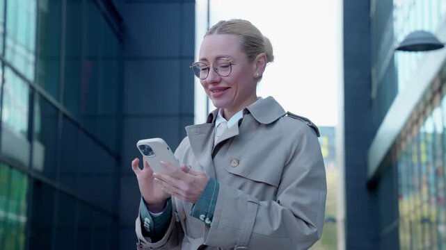 Woman checks her phone for messages while standing outside a tall building on a clear day in the city