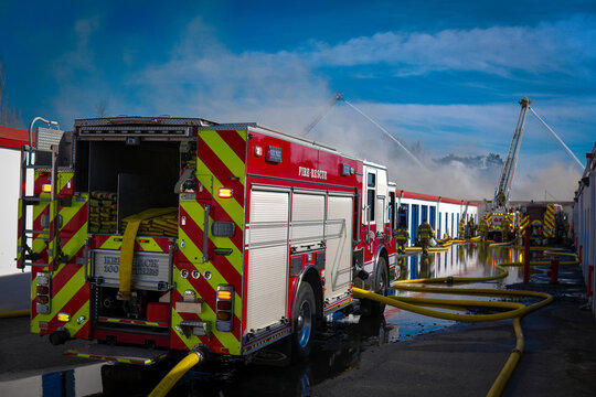 Fire engine in the foreground with supply hose delivering water to aerial apparatus in the background spraying water on a burning storage facility as smoke rises into a blue sky.  