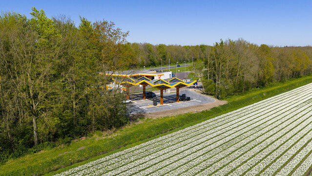 Bant, Netherlands - 21 April 2026: Aerial view of the Fastned charging station with its iconic yellow solar roofs situated between a vibrant white tulip field and a lush green forest.
