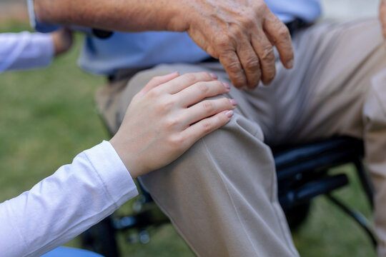 Hand resting gently on knee of elderly senior man in wheelchair showing caregiver support touch care and deep compassion providing comfort and empathy for vulnerable patient outdoors in park