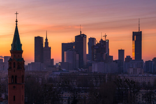 Warsaw, Poland - 05 March 2026: Aerial view of the Warsaw skyline with modern skyscrapers silhouetted against a vibrant sunset sky, featuring a historic church spire in the foreground.