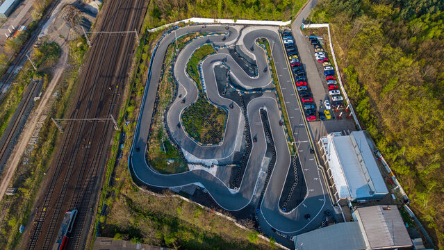 Prague, Czechia - 21 April 2026: Aerial view of KartPlanet go-kart track with racing karts on the circuit, a parking area filled with cars, and adjacent railway tracks surrounded by green trees.