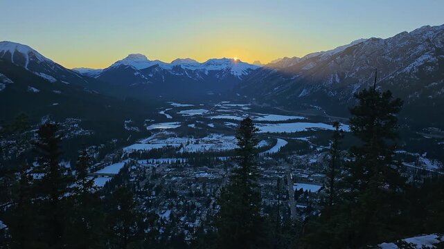 Sun setting over Banff, Alberta, with snowy Bow Valley, mountain town below, dark evergreen silhouettes, glowing horizon, frozen river curves, and dramatic winter Rockies scenery.