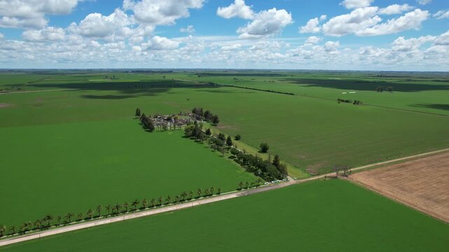 Drone flyover at altitude revealing a small town cemetery among fertile industrial soybean corn and alfalfa plantations under a cloudy sky in the Argentine agroindustrial region
