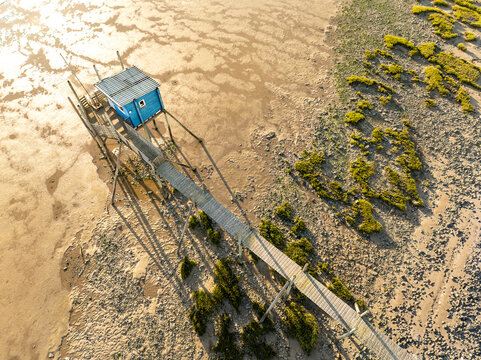Yves, France - 22 September 2023: Aerial view of a traditional blue fishing hut on stilts with a long wooden walkway over tidal flats and green vegetation during golden hour.