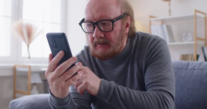 Elderly man wearing glasses sitting on sofa and concentrating on using smartphone. Senior male learning modern technology, surfing internet, scrolling social media and having fun during online time.