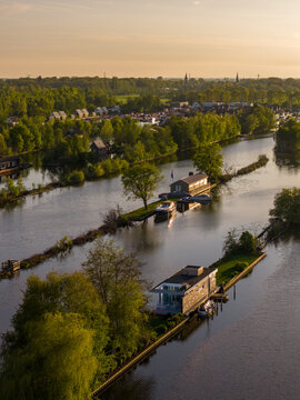 Breukelen, Netherlands - 25 April 2026: Aerial view of Scheendijk recreational area featuring narrow islands with modern houses and boats surrounded by lush trees during golden hour.