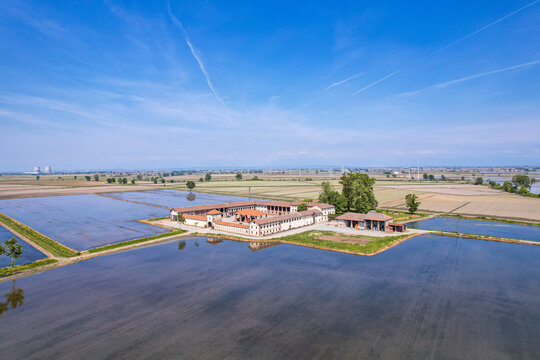 Aerial view of a traditional farm complex surrounded by flooded rice fields under a clear blue sky in Trino, Piedmont, Italy.