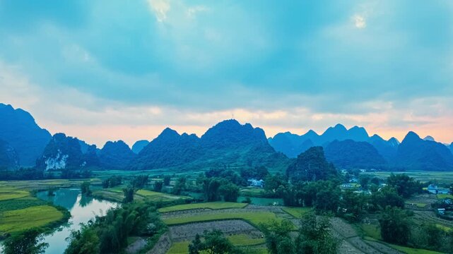 A breathtaking Aerial view of panoramic view of karst mountains, winding river, and rural farmland under a dramatic cloudy sky during sunrise. Calm Waterway Through Farmland