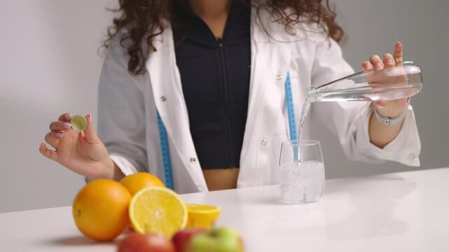 Waist up shot of young dietitian standing behind table with apples and oranges, pouring water from a clear glass bottle into a glass and closing it. Healthy hydration and nutrition concept on white ba